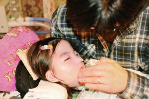 『穂花(ほのか)』メイン写真。 お父さん・秀勝さんがベッドの上の娘・穂花さんを抱き、口元に触れている。目を瞑ったままの穂花さんですが、花柄のピンで髪を止め、ベッドにはハート型の「I LOVE YOU」と書かれたクッションが。生活に溢れる愛情が伝わってきます。