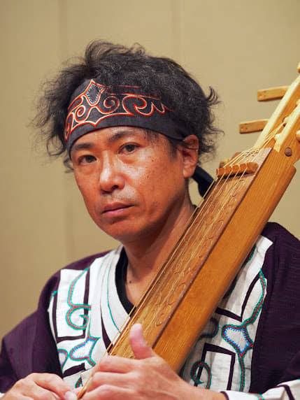 千葉伸彦さん画像。アイヌの衣装を着て、頭にはマタンプシを巻いている。木製の弦楽器、トンコリを抱えている。